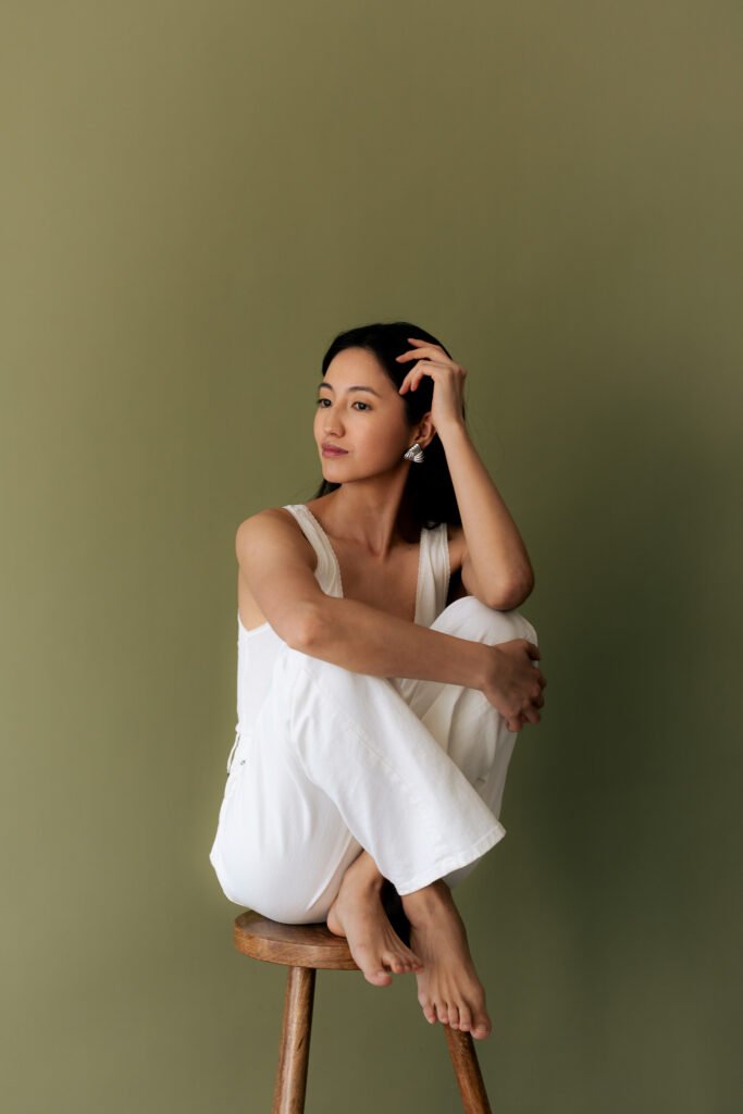 Woman in white outfit seated on wooden stool against olive green background with hand in hair representing personal brand identity